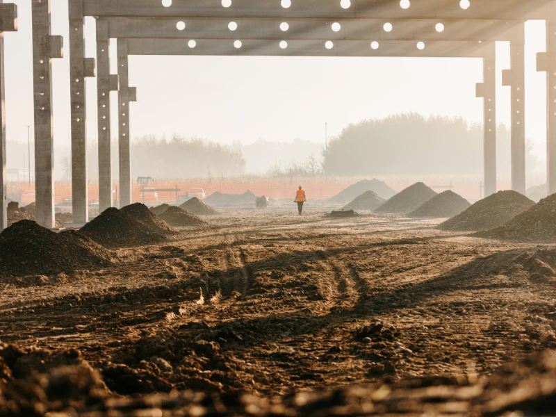 Worker in orange vest walks across muddy construction site with dirt mounds, framed by concrete pillars & misty sun.
