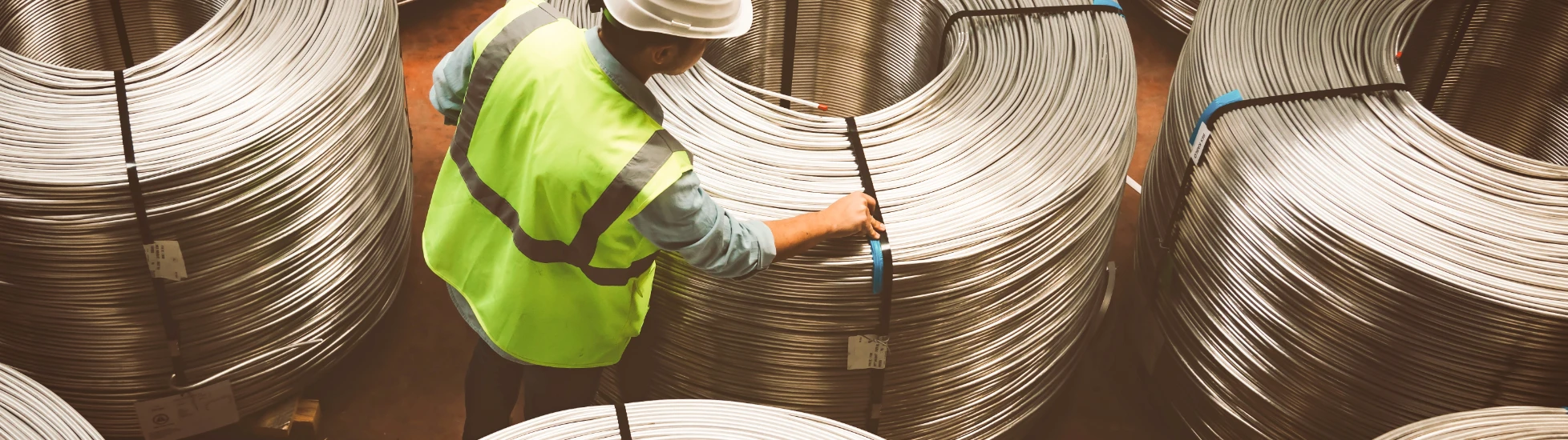 Overhead: Industrial worker (white hard hat, yellow-green vest) examines a large spool of metal wire/rebar in a warehouse.