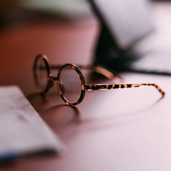 A pair of round, tortoise-shell eyeglasses rests on a desk next to a sheet of paper, with a soft, warm light casting a shallow depth of field.